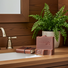 Load image into Gallery viewer, A rustic bar of soap from 'Mammy's Soap Co.' sits on a wooden bathroom counter next to a brass faucet, a small leather notebook, and a potted fern.
