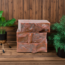 Load image into Gallery viewer, Three stacked rectangular bars of handmade soap with a swirled terracotta and brown pattern, displayed on a wooden surface against a wooden plank wall, flanked by green potted plants.
