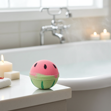 Load image into Gallery viewer, Watermelon-shaped bath bomb on a bathtub ledge with candles and soap.
